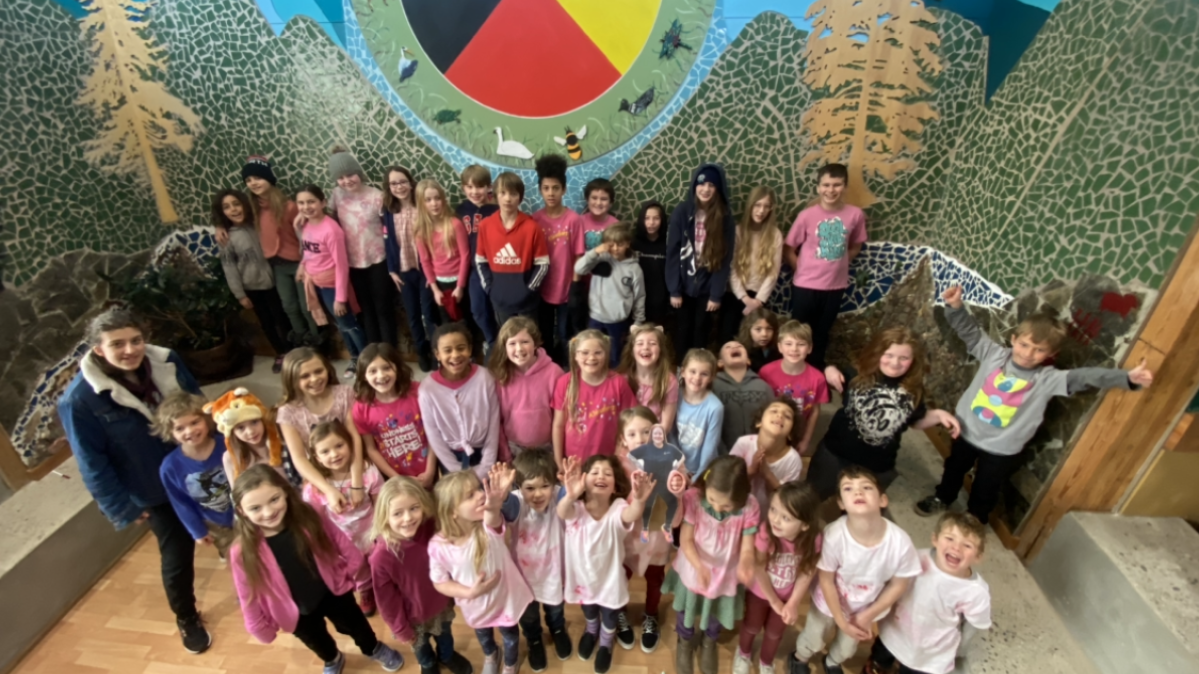 Birdseye view of elementary school class in front of a forest mural at the centre of which is a painting of a coloured circle depicting the First Nations' four directions.