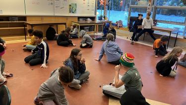 Over a dozen students seated facing one another in pairs, on a classroom floor.