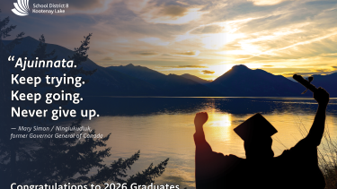 A silhouette of a student in cap and gown standing in front of Kootenay Lake at sunset